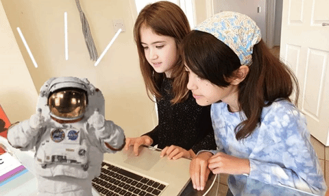 Two young girls with long brown hair sat facing a laptop at a table. In the corner of the image an animation of of a astronaut is giving a double thumbs up. 